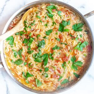 a pan with cooked orzo, tomatoes, parsley, and a wooden spoon.