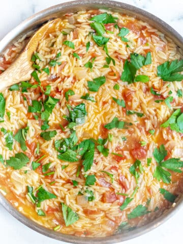 a pan with cooked orzo, tomatoes, parsley, and a wooden spoon.