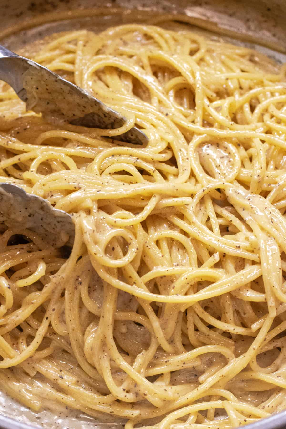 a pan with cooked spaghetti and pepper with tongs.