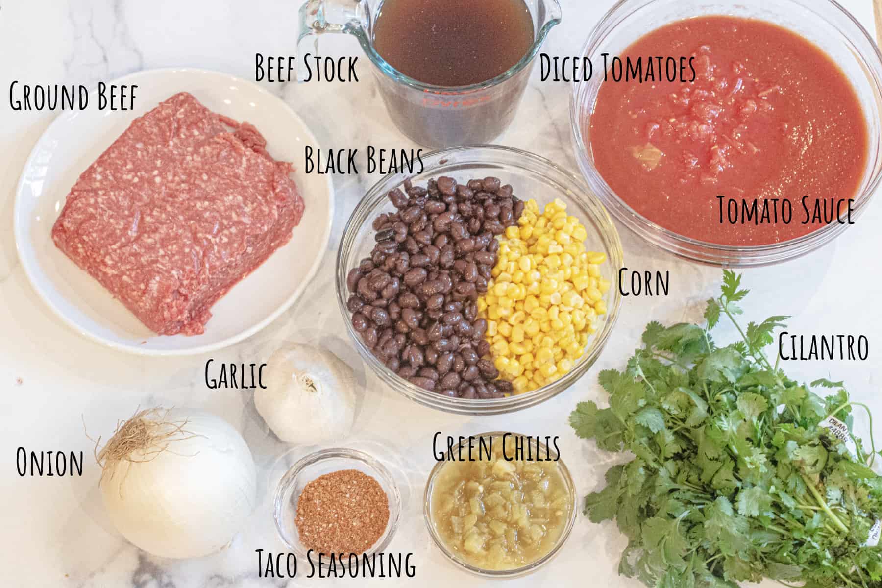 raw ground beef, corn, beans, beef stock, a bowl of tomato paste, and diced tomatoes, onion, garlic, slices, diced green chilis, and cilantro all labeled.
