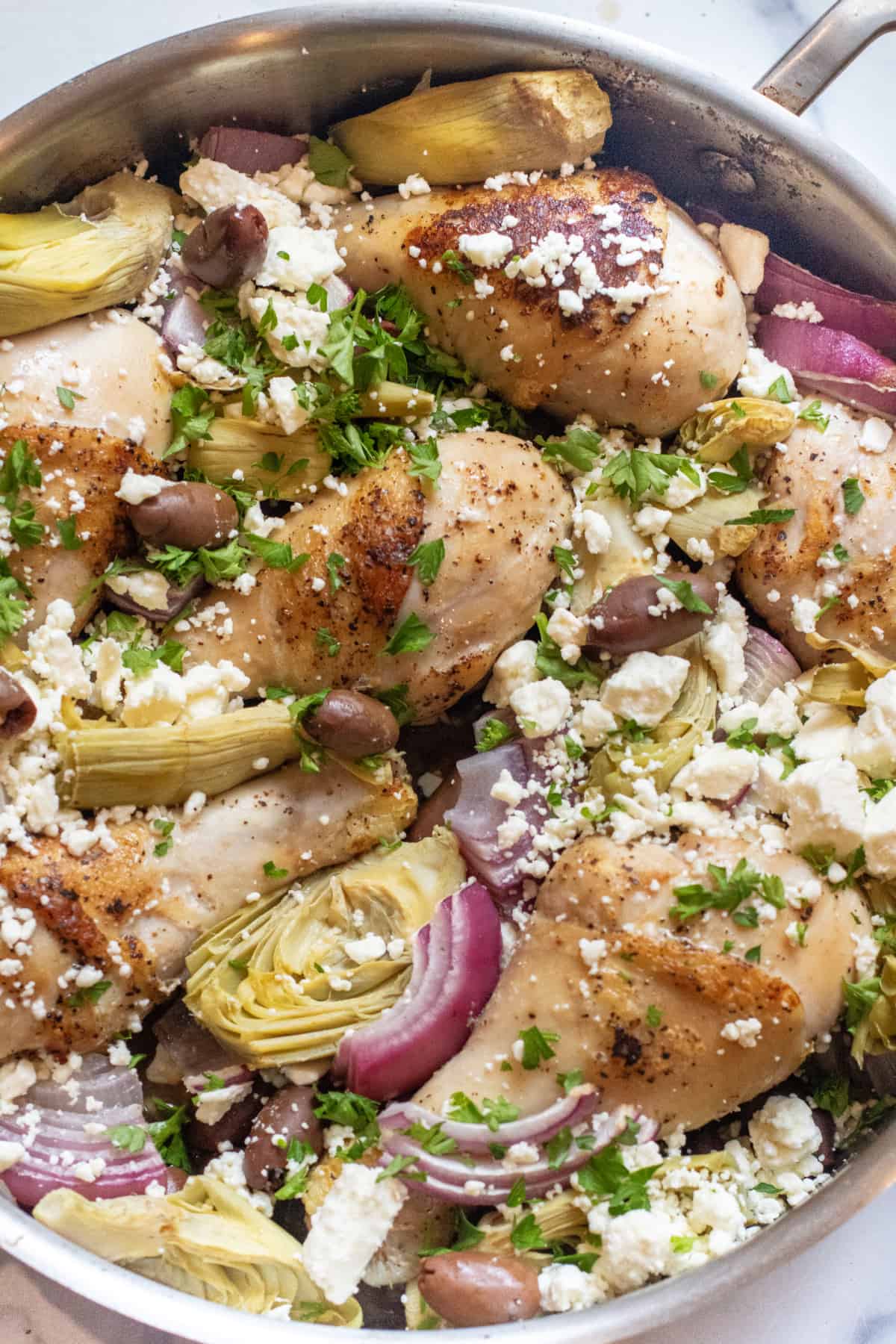 a pan of cooked chicken legs with feta, olives, onions, and artichokes.