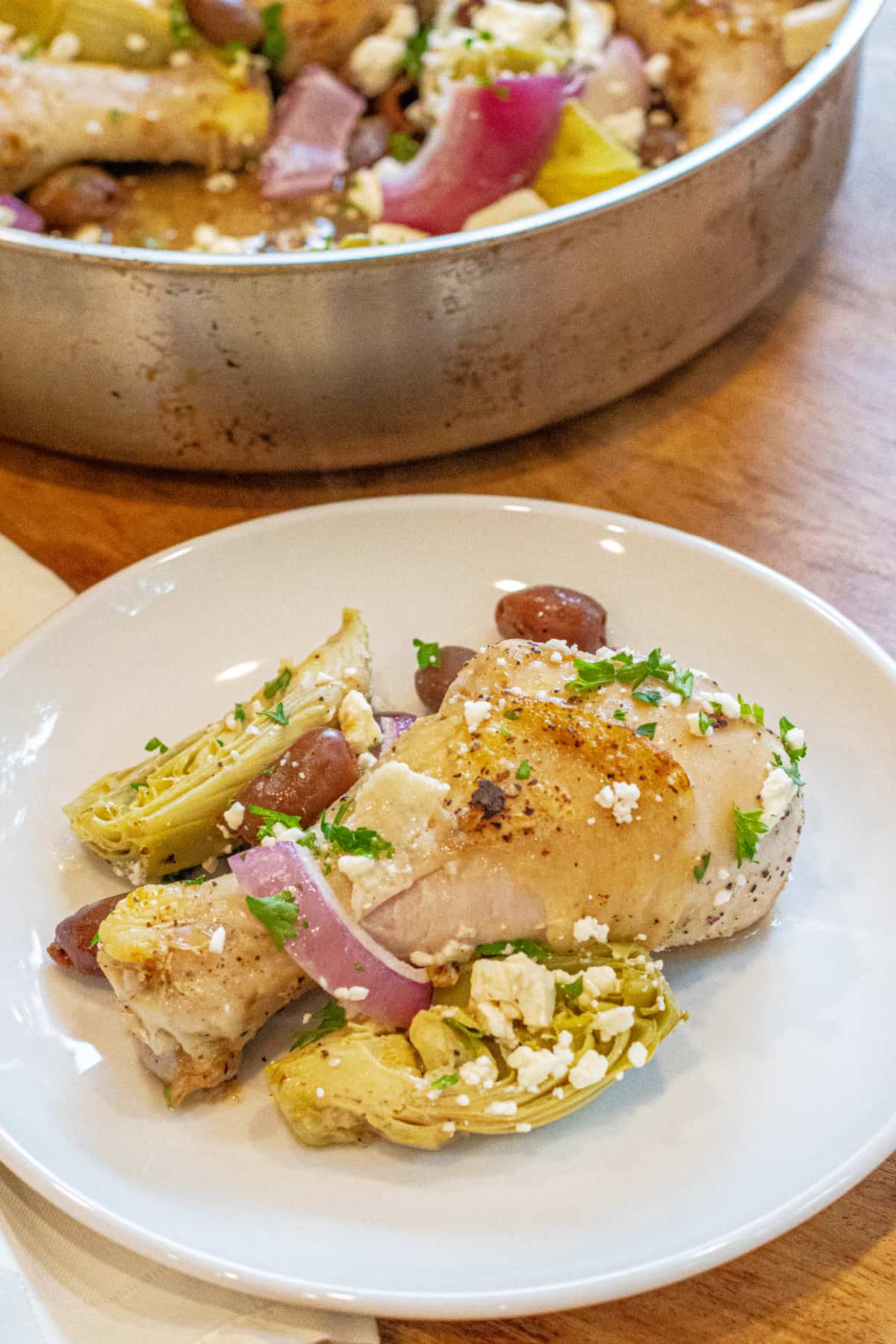 a plate with a cooked chicken leg and olive and onions, artichokes, and feta.