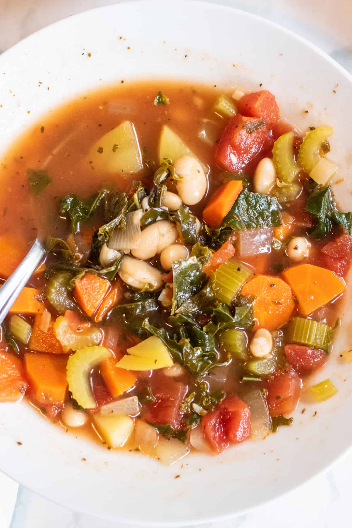 an overhead of a bowl of soup with kale, potatoes, beans, and carrots with a spoon in it.