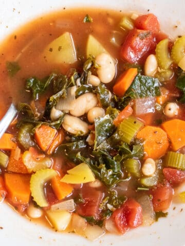 an overhead of a bowl of soup with kale, potatoes, beans, and carrots with a spoon in it.