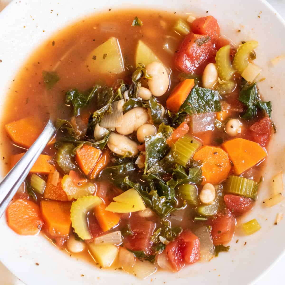 an overhead of a bowl of soup with kale, potatoes, beans, and carrots with a spoon in it.