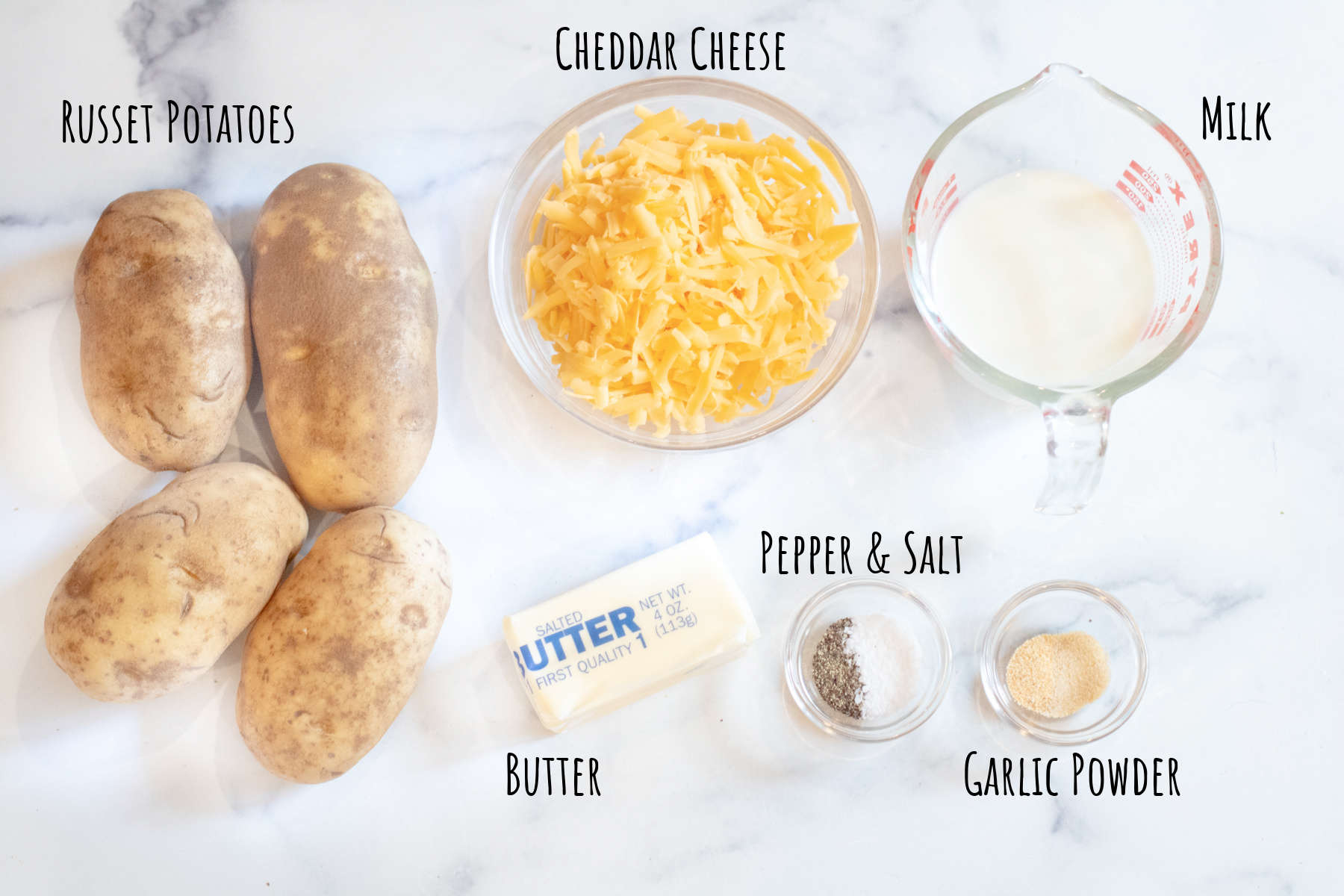 potatoes, a bowl of cheese, and a bowl of flour with butter, salt and pepper, and garlic powder on labeled on a counter.