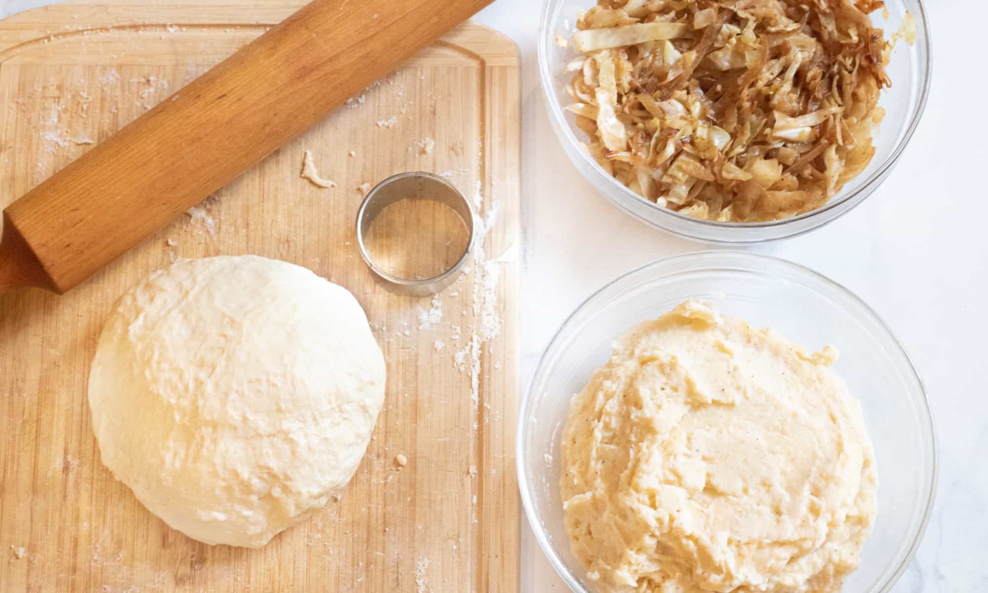 a cutting board of rolling pin and a ball of dough and then a bowl of cabbage and a bowl of mashed potatoes.