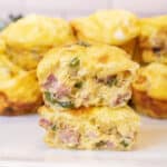 two halves of egg cups stuffed with ham, peppers, and onion stacked on a plate of other egg cups.