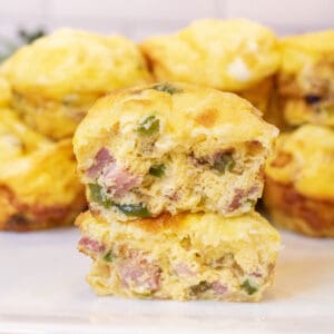 two halves of egg cups stuffed with ham, peppers, and onion stacked on a plate of other egg cups.