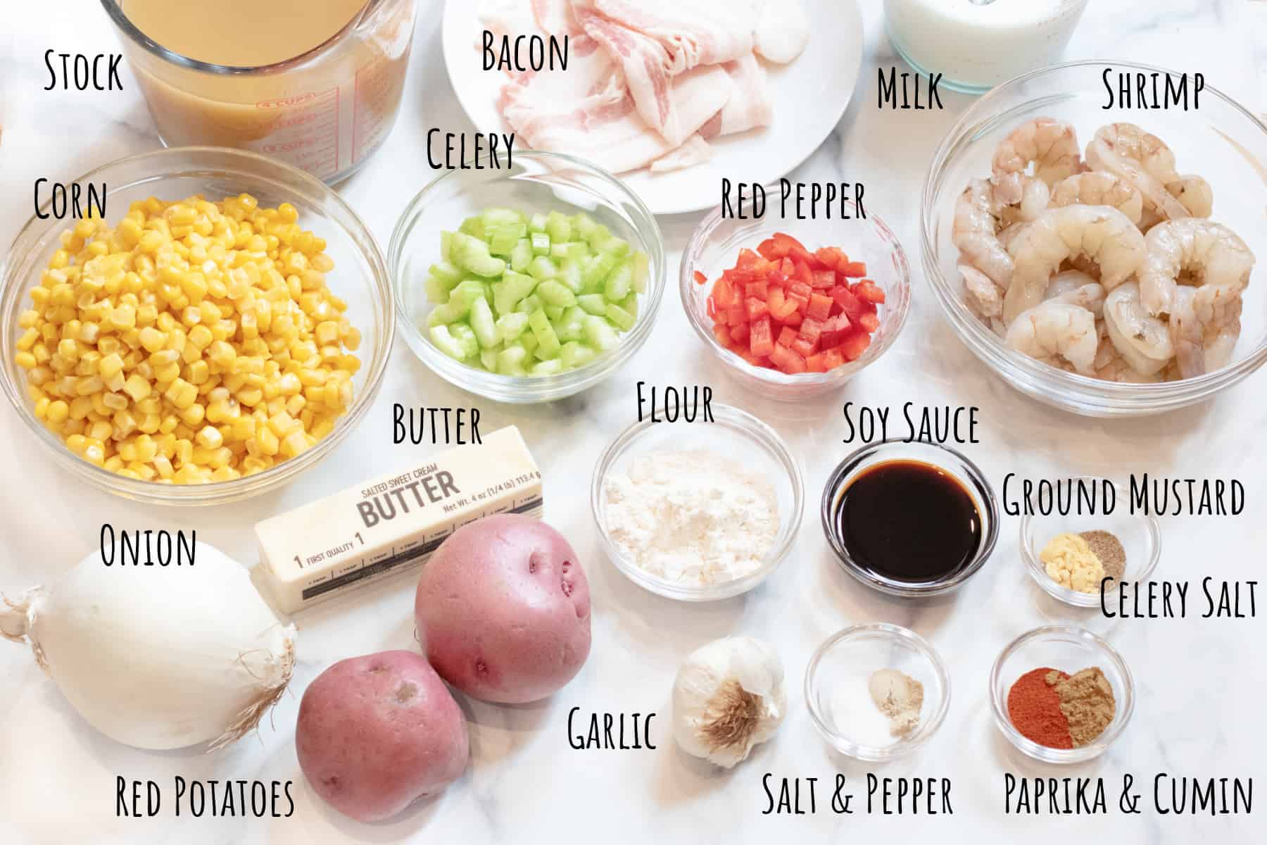 corn, shrimp, stock, milk, raw bacon, potatoes, onions, celery, red pepper, flour, soy sauce, spices and garlic on a counter labeled.