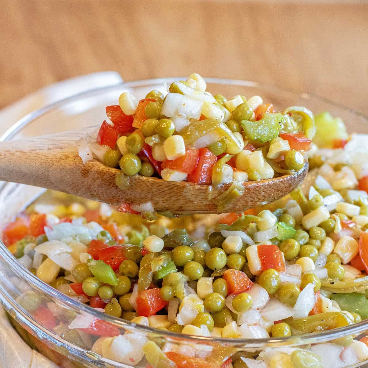 a wooden spoon holding up some vegetable salad with corn, peas, red pepper, and onion.