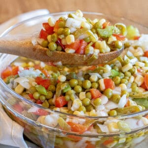 a wooden spoon holding up some vegetable salad with corn, peas, red pepper, and onion.