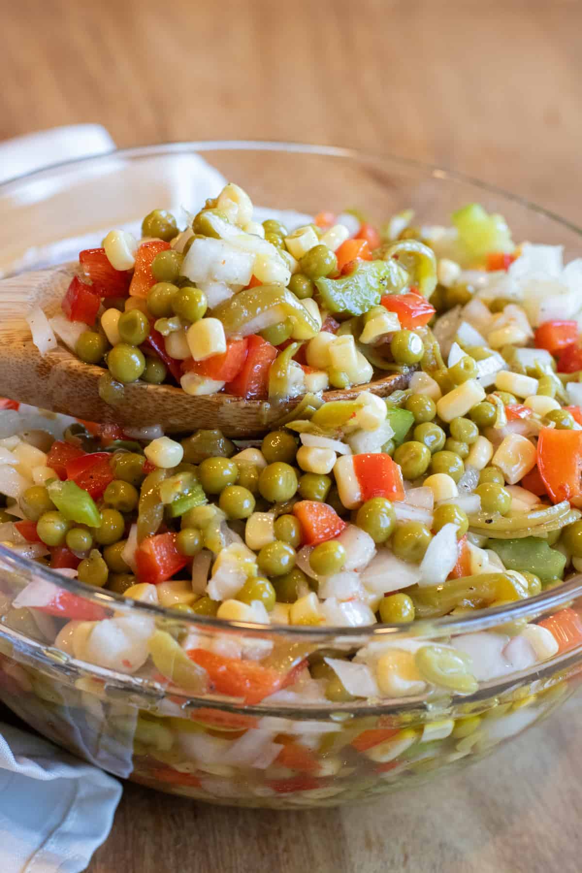a wooden spoon holding up some vegetable salad with corn, peas, red pepper, and onion.