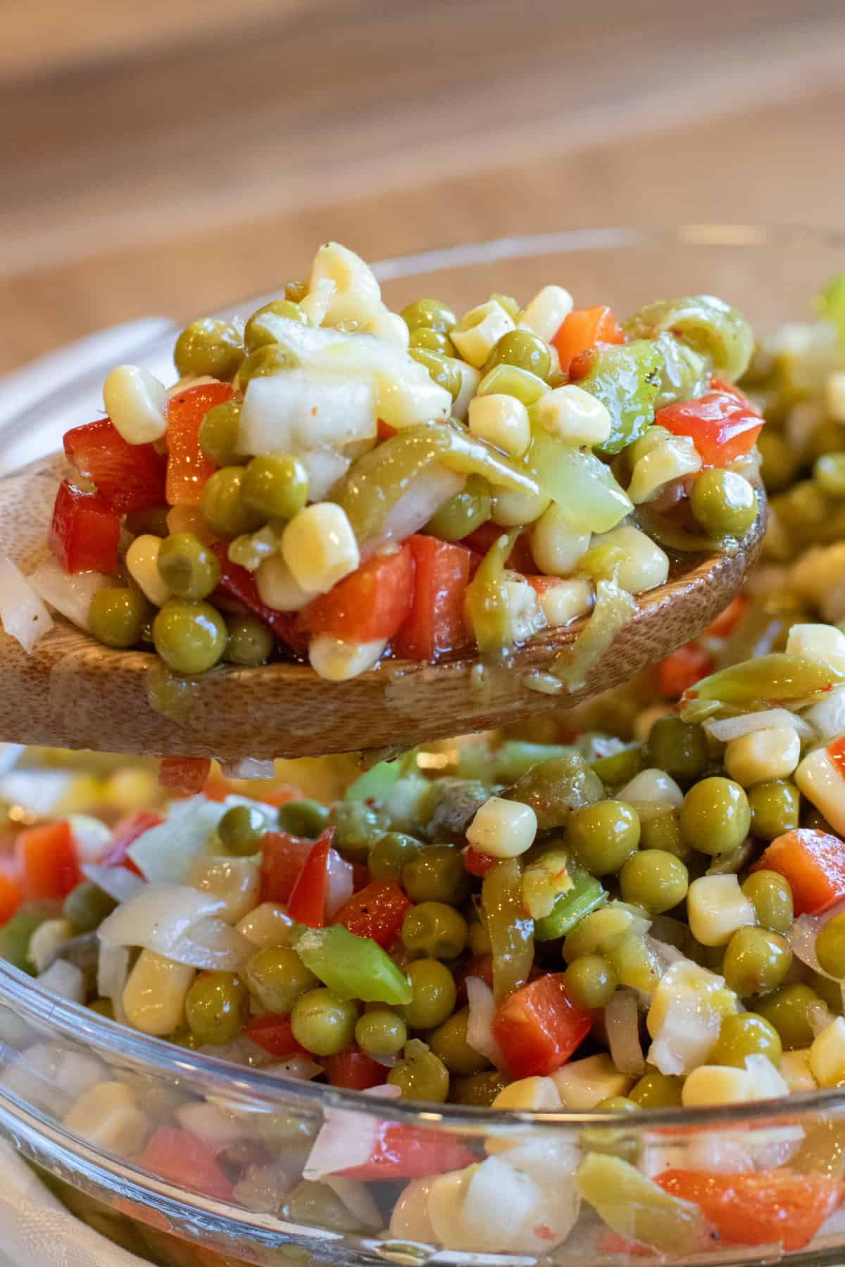a wooden spoon holding up some vegetable salad with corn, peas, red pepper, and onion.