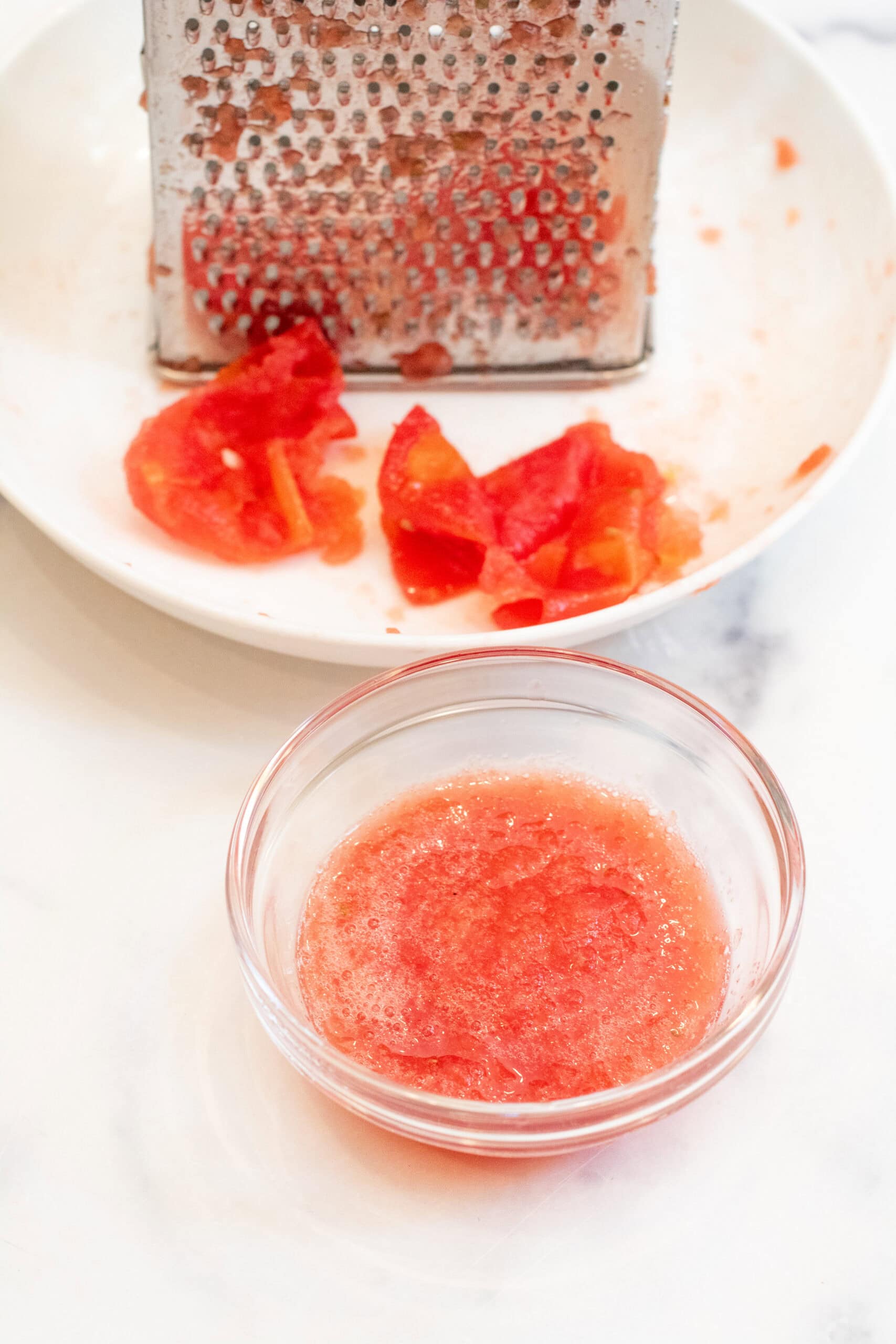 a bowl of tomato sauce in front of a grater on a plate and tomato skins.