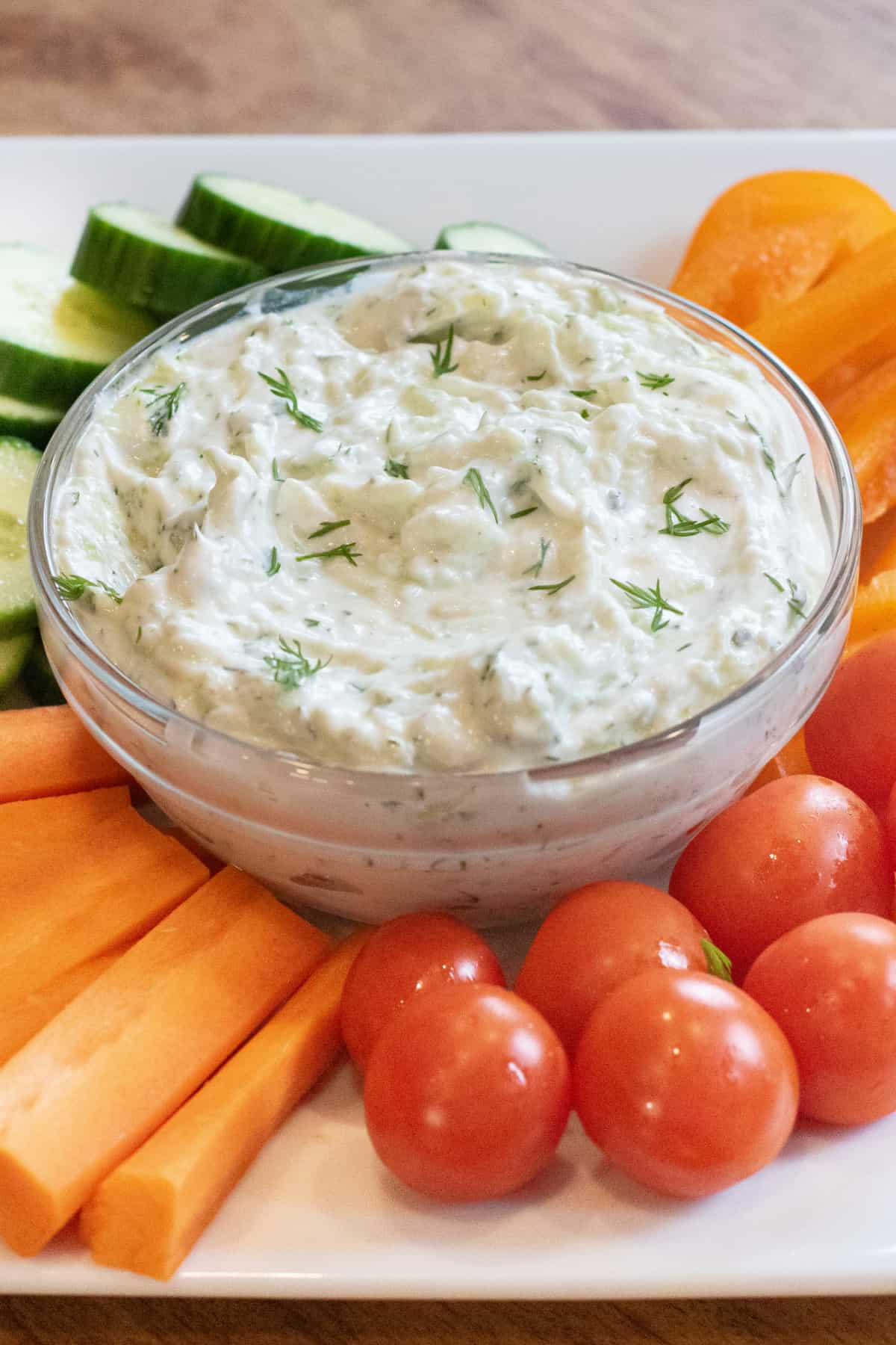 an overhead of a bowl of dill tzatziki surrounded by cucumbers, carrots, peppers, and tomatoes.