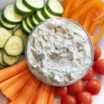 an overhead of a bowl of dill tzatziki surrounded by cucumbers, carrots, peppers, and tomatoes.