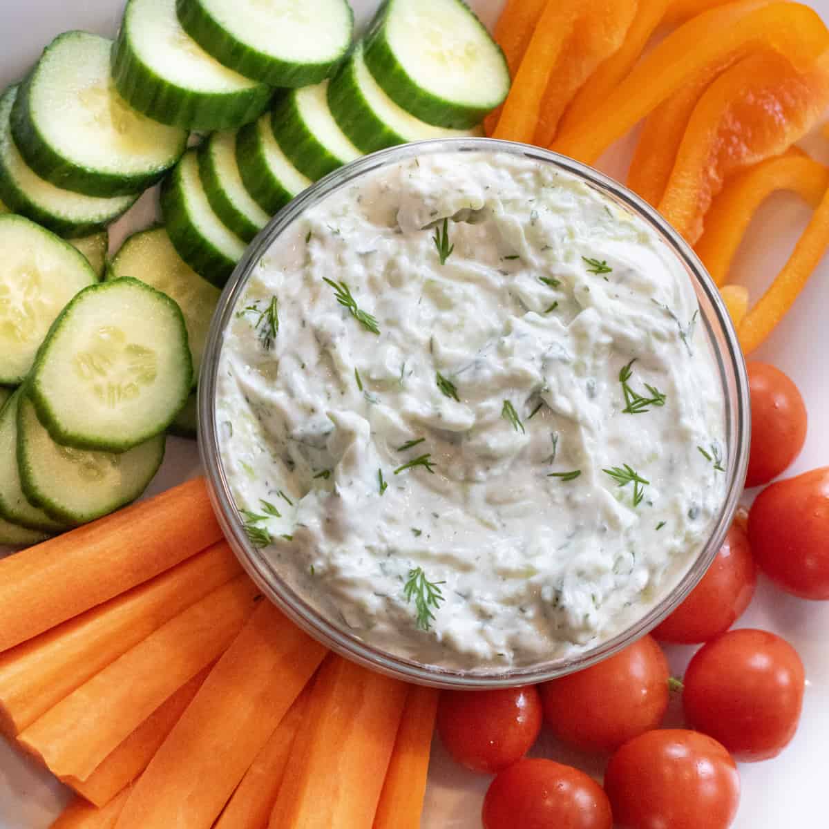 an overhead of a bowl of dill tzatziki surrounded by cucumbers, carrots, peppers, and tomatoes.