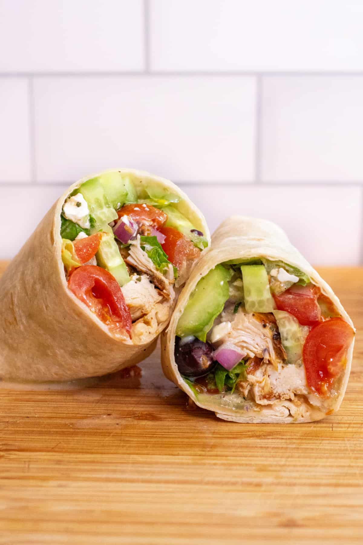 a halved chicken and greek salad wrap on a cutting board.