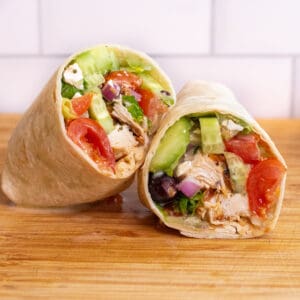 a halved chicken and greek salad wrap on a cutting board.