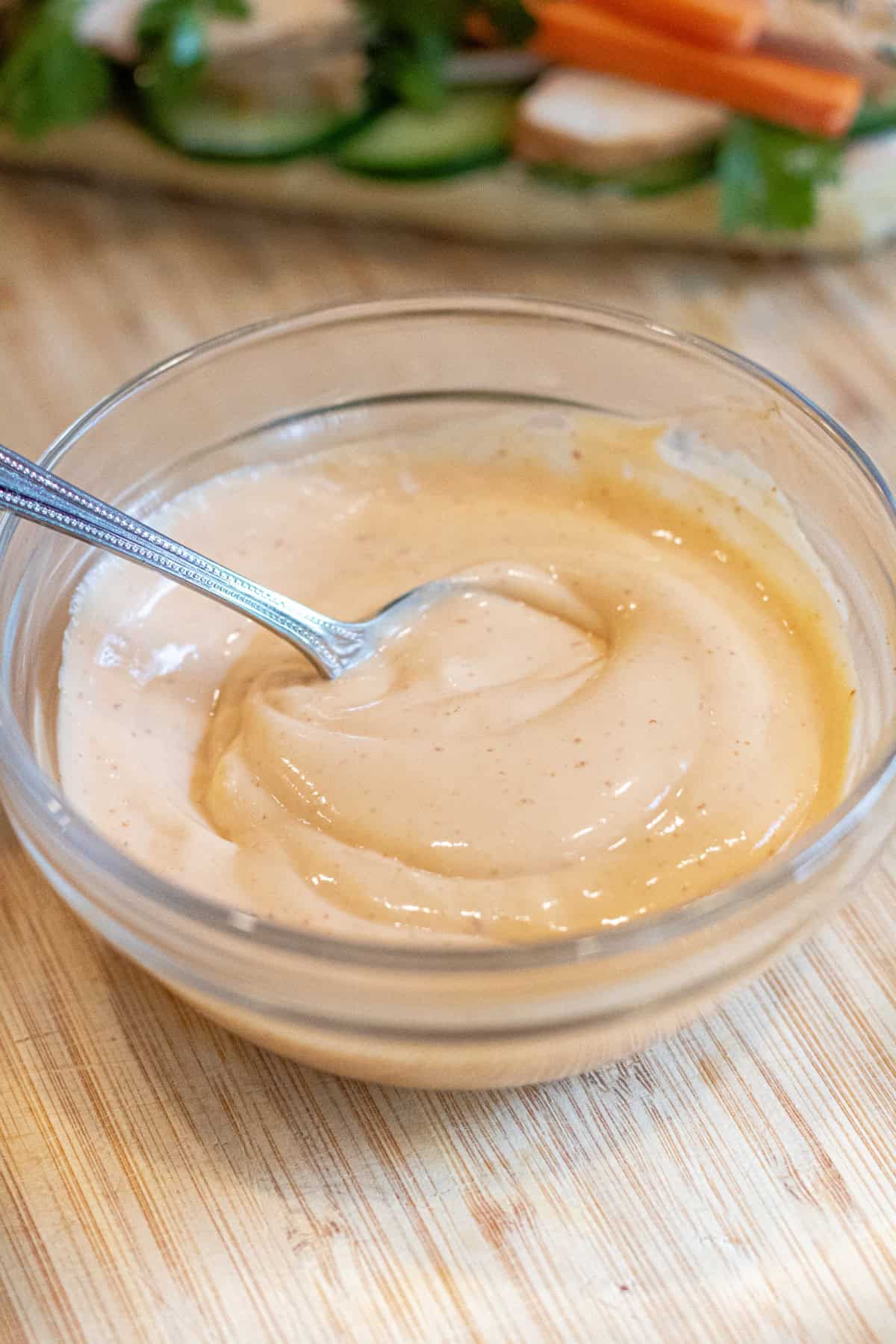 a bowl of mixed sriracha mayo with a spoon on a cutting board in front of sandwich with cucumber.