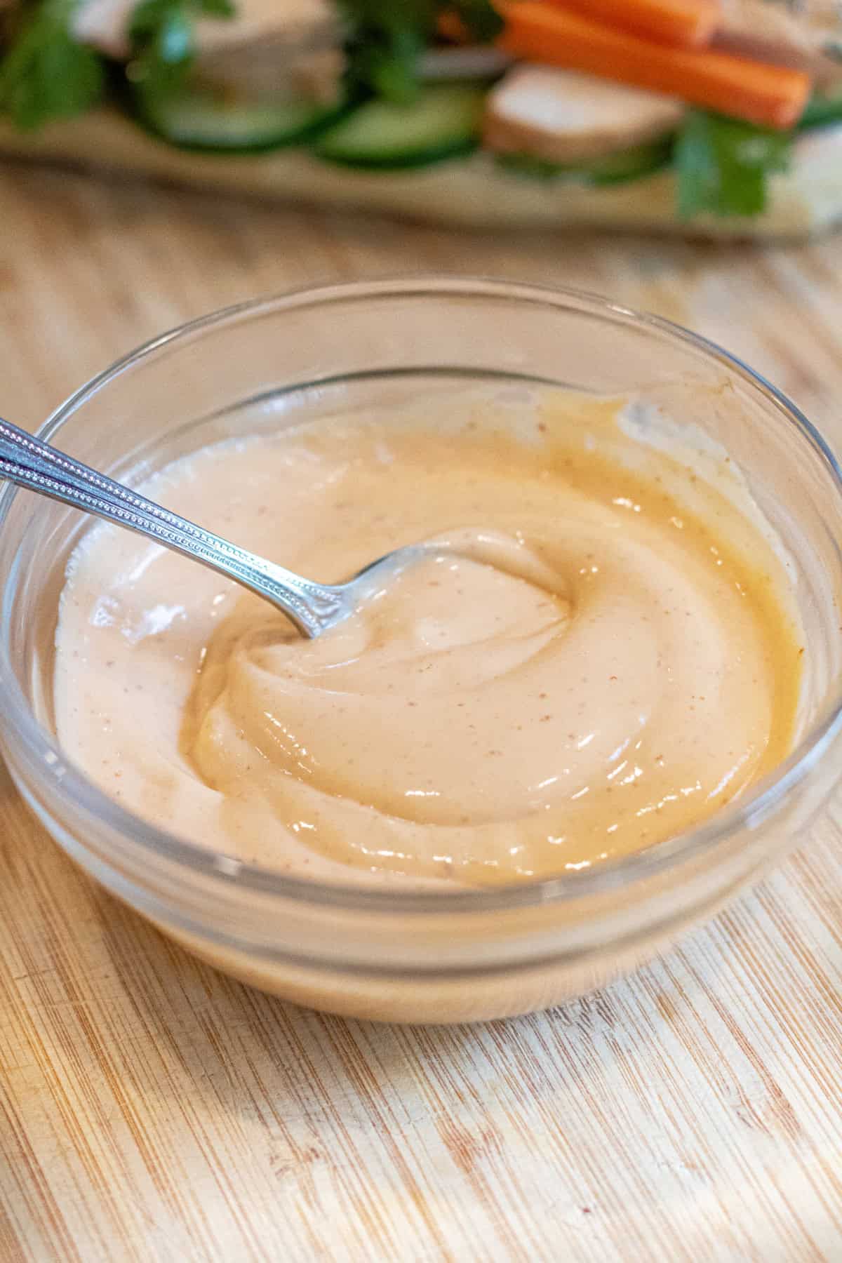 a bowl of mixed sriracha mayo with a spoon on a cutting board in front of sandwich with cucumber.