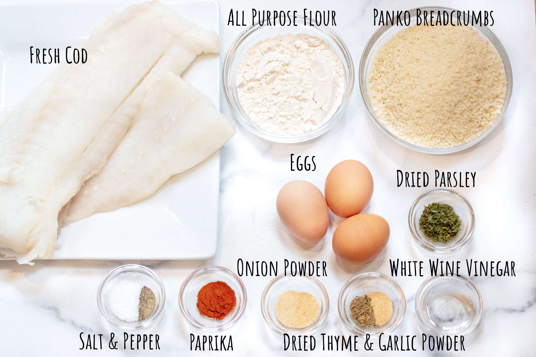 fresh cod, bowls of flour, breadcrumbs, eggs, herbs and spices labeled on a counter.