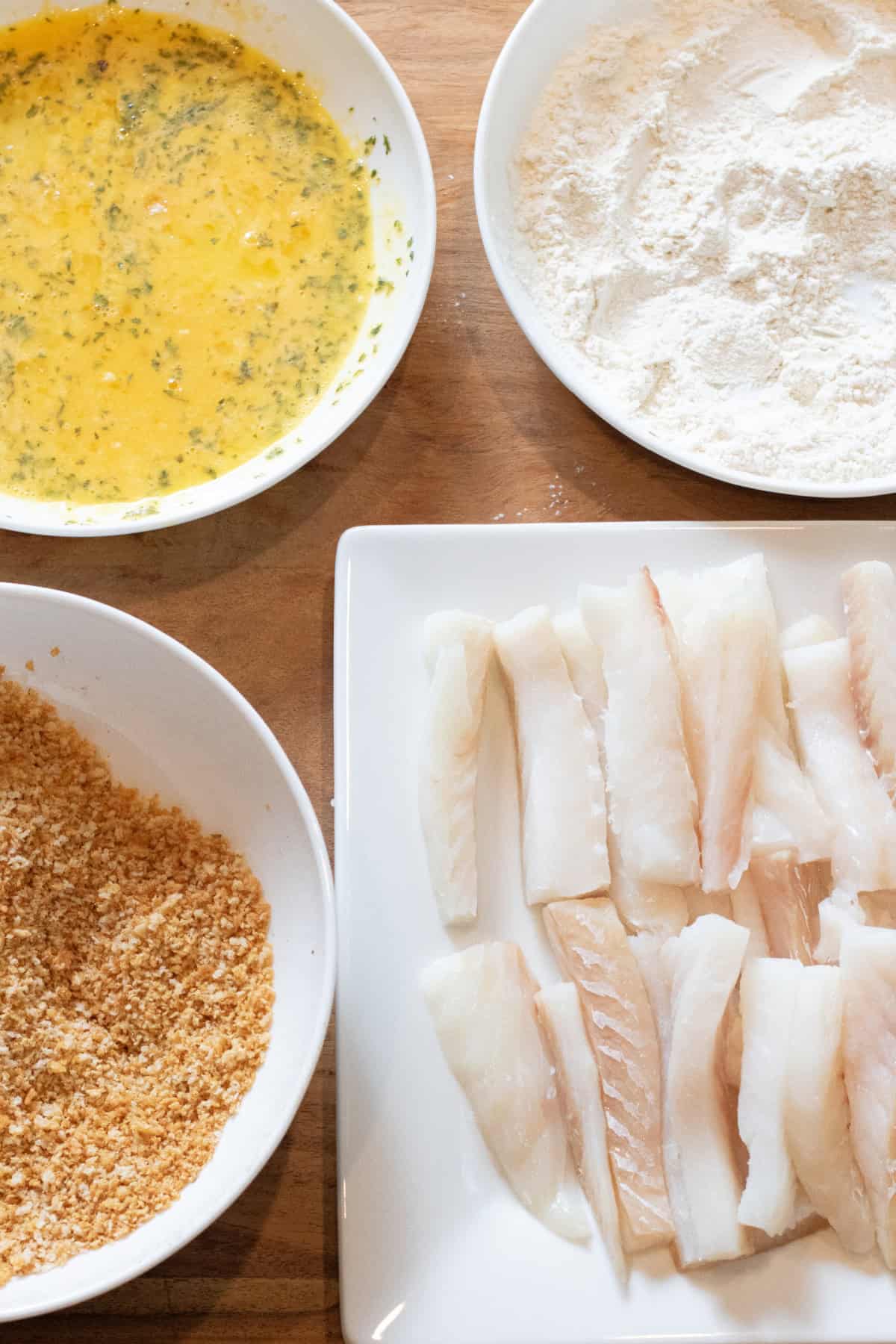 bowls of toasted bread crumbs, egg wash, and flour and a plate of cut cod in sticks.