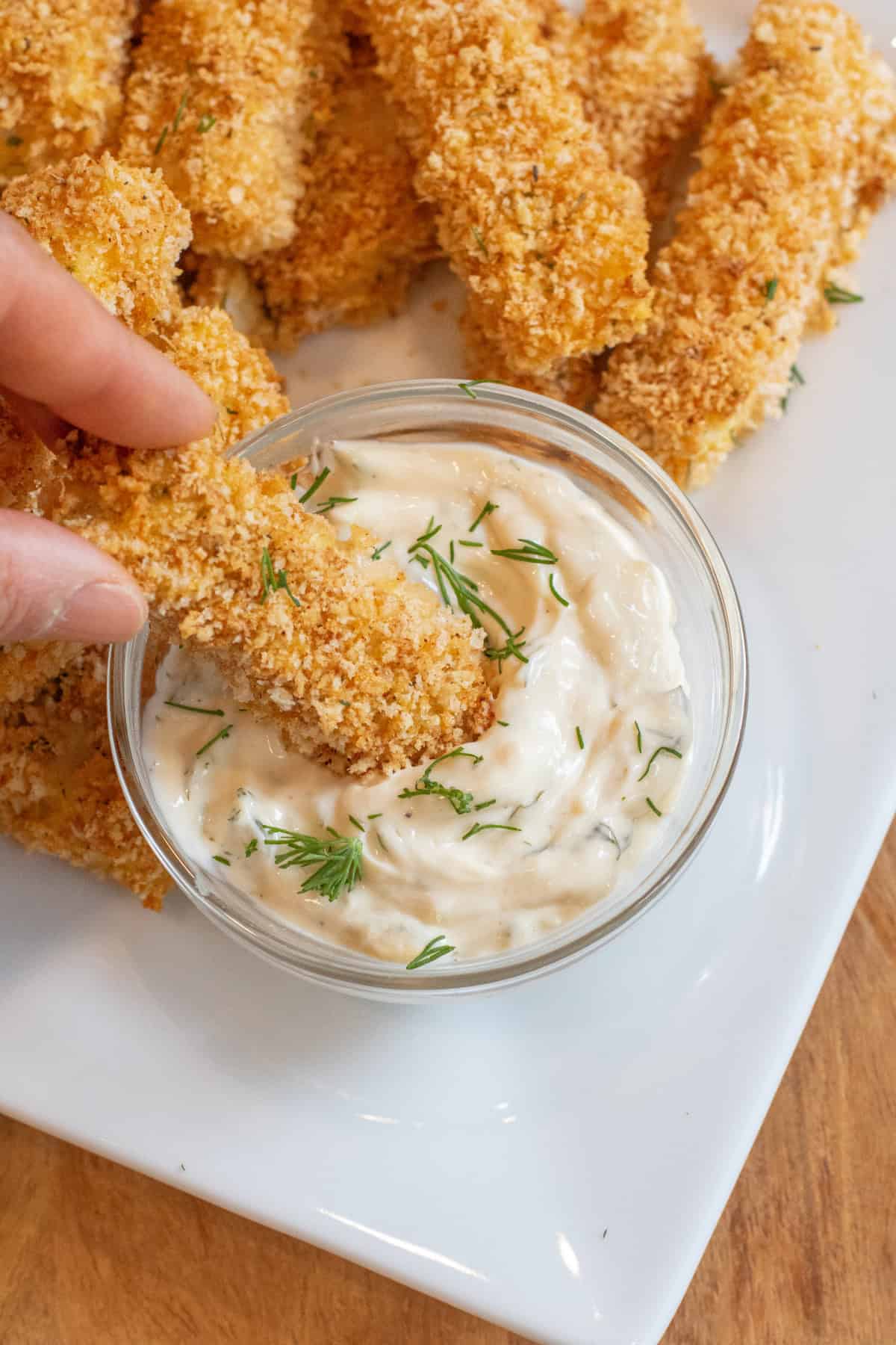 a hand dipping a breaded fish stick into a bowl of tartar sauce.