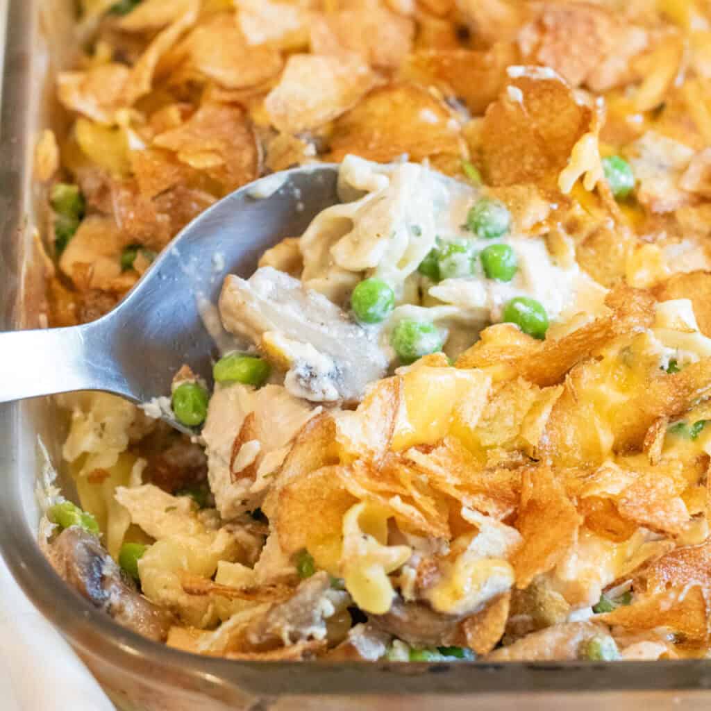 a spoon in a casserole with potato chips and peas, mushrooms, noodles, and tuna.