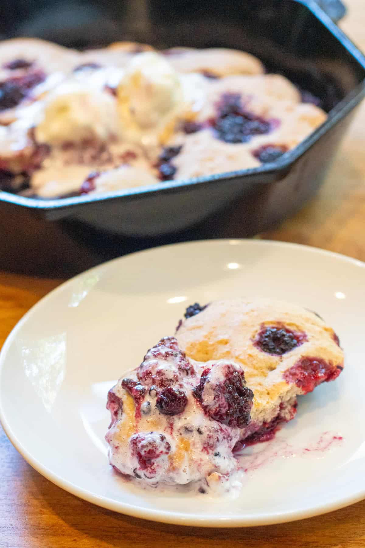 a slice of a cobbler with ice cream and berries in front of a cast iron skillet with cobbler.