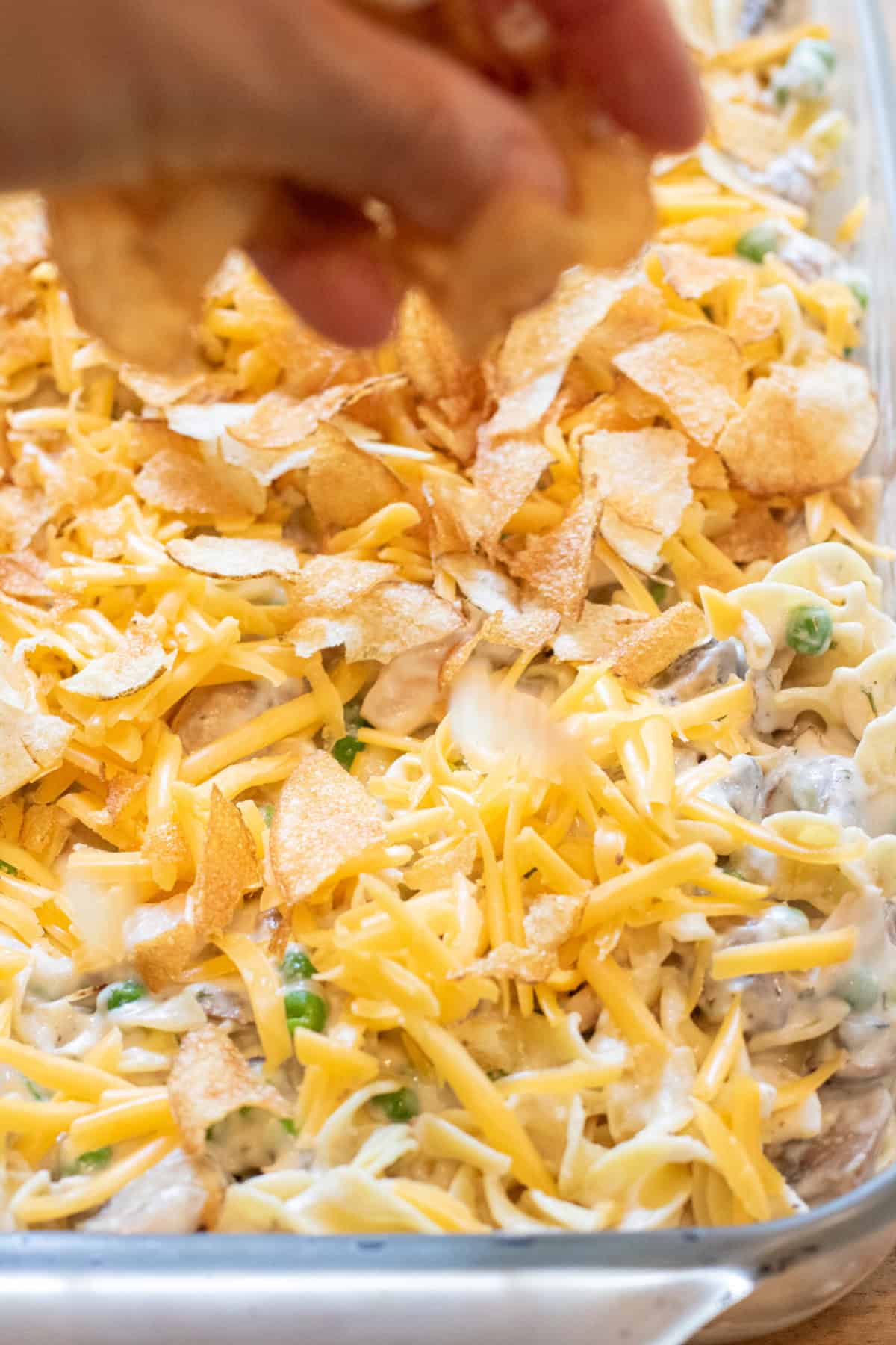 a hand adding crushed potato chips on a top of a cheddar cheese, peas, tuna, mushroom soup casserole.
