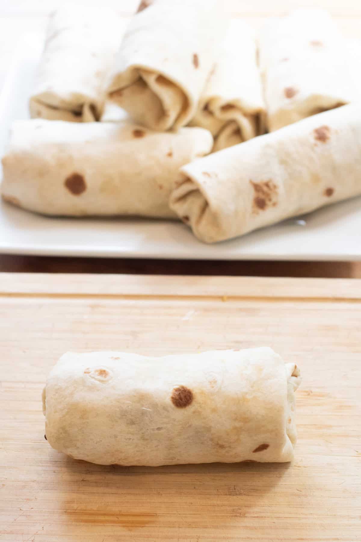 a rolled burrito on a cutting board and then a plate of burritos behind it.