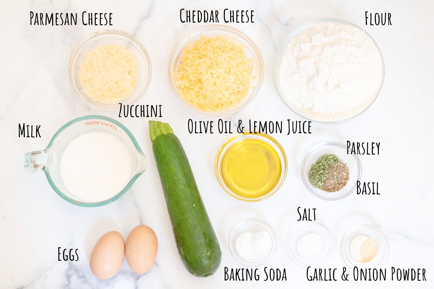 zucchini, cheddar cheese, parmesan cheese, flour, milk, olive oil and lemon juice, eggs, herbs, salt, and baking powder and baking soda in bowls labeled.