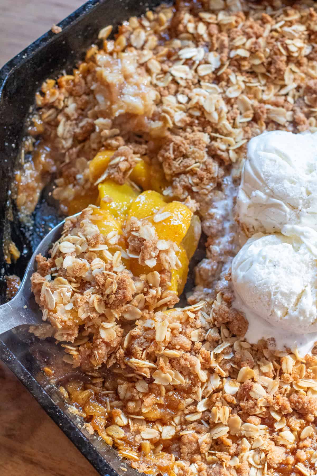 a cast iron of baked peach crisp with two scoops of vanilla ice cream and a spoon scooping some out.