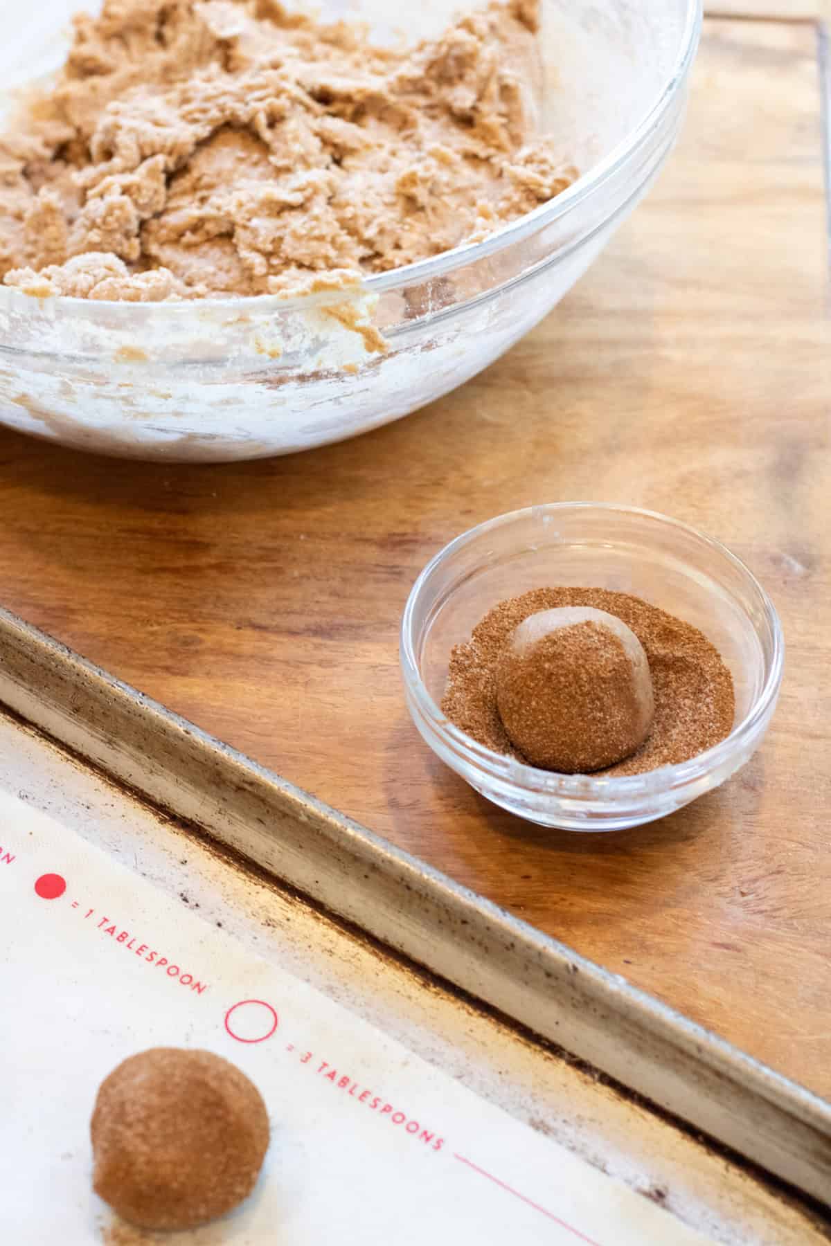 a bowl of dough with a dough ball being rolled in a bowl of spices next to a baking tray.
