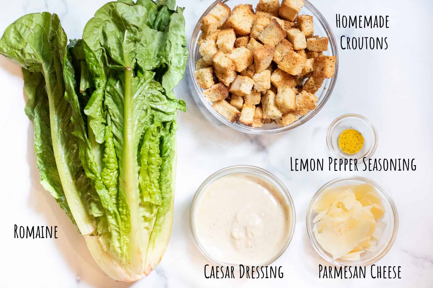 a head of lettuce and a bowl of croutons, caesar dressing, shaved parmesan cheese, and lemon pepper seasoning all labeled.