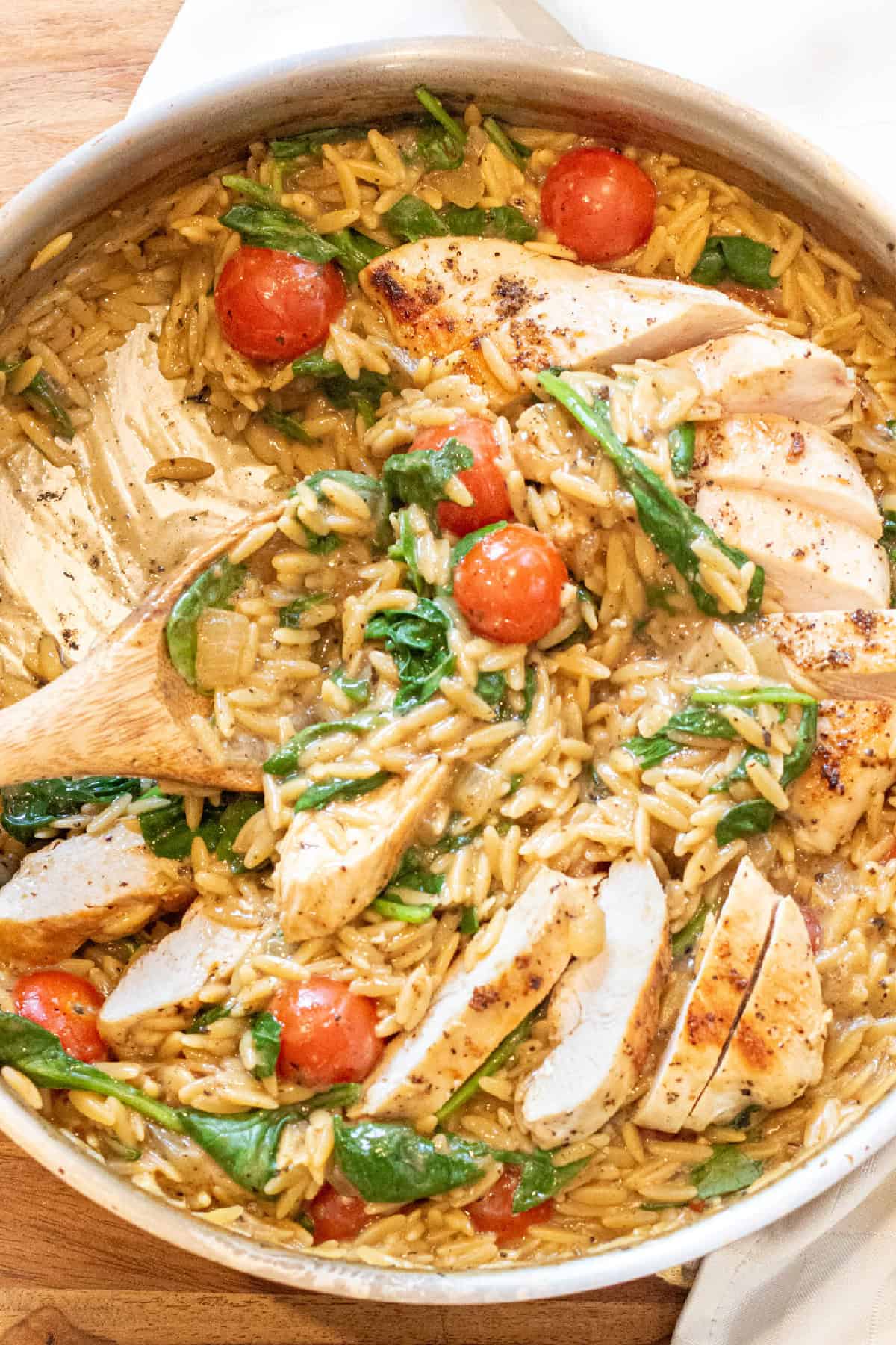 an overhead of a skillet with cooked sliced chicken and orzo and spinach and tomatoes with a wooden spoon.