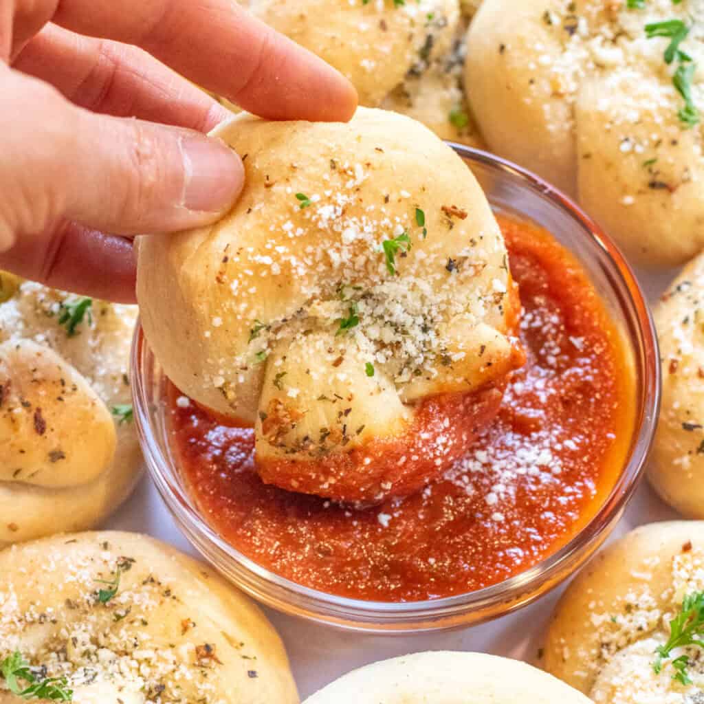 a hand dipping a garlic knot into a bowl of marinara sauce.