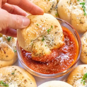 a hand dipping a garlic knot into a bowl of marinara sauce.