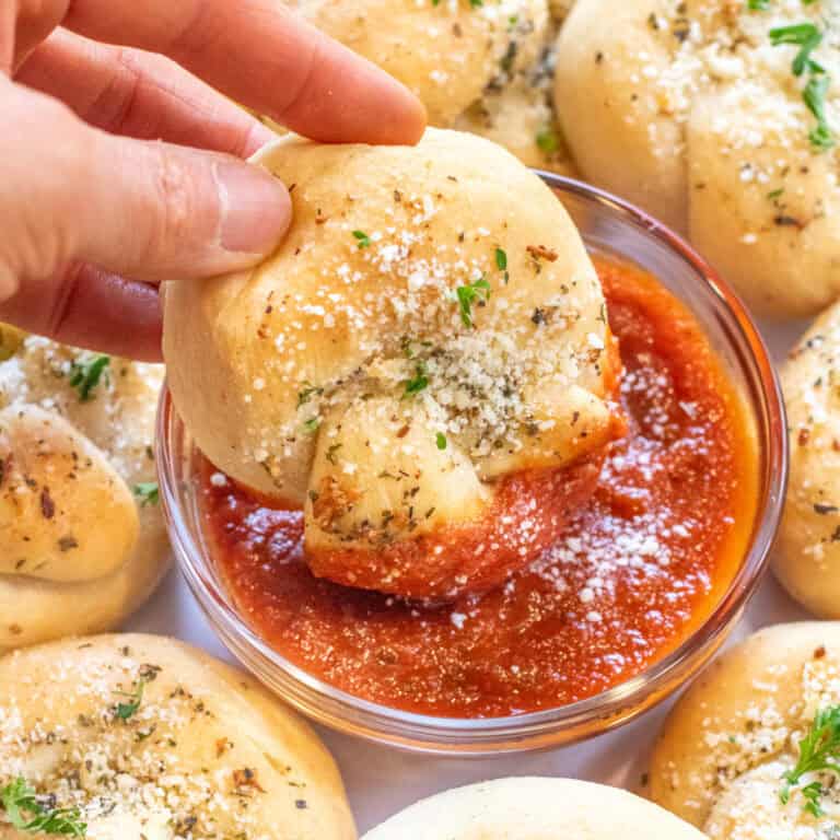 a hand dipping a garlic knot into a bowl of marinara sauce.