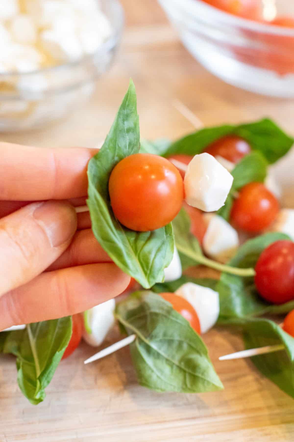 a hand holding a toothpick of a basil leaf, cherry tomato, and a mozzarella pearl.