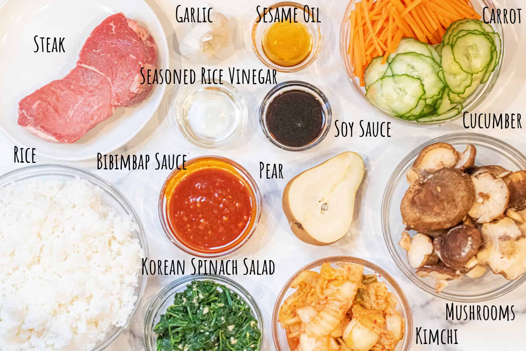 sirloin, a bowl of rice, sauce, kimchi, korean spinach salad, shiitake mushrooms, cucumbers, carrots, soy, half a pear, garlic, sesame oil, and vinegar and soy sauce labeled.