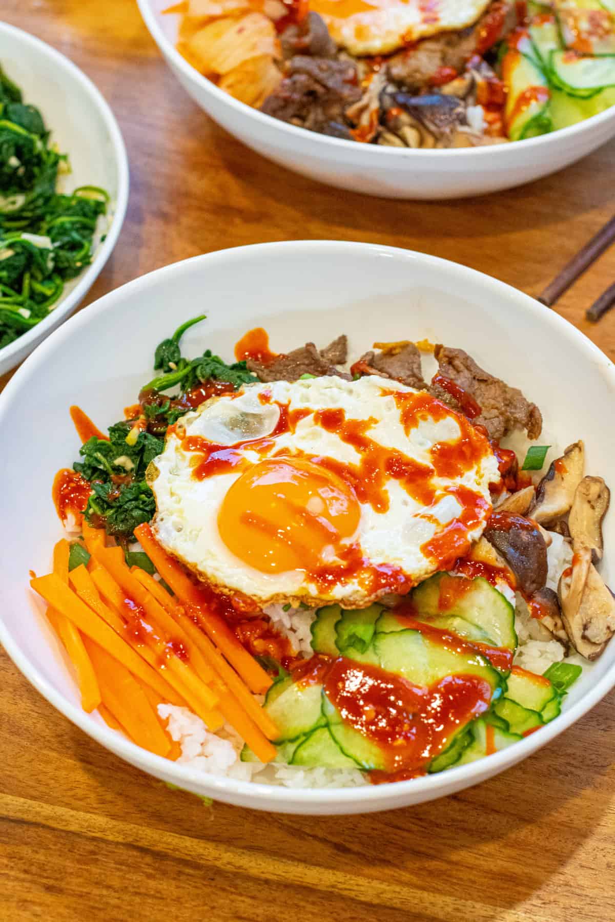 overhead of a big wide bowl of sliced carrots, cucumbers, spinach salad, beef, mushrooms, and an egg on top with sauce.