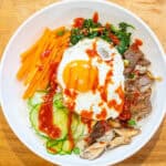 overhead of a big wide bowl of sliced carrots, cucumbers, spinach salad, beef, mushrooms, and an egg on top with sauce.