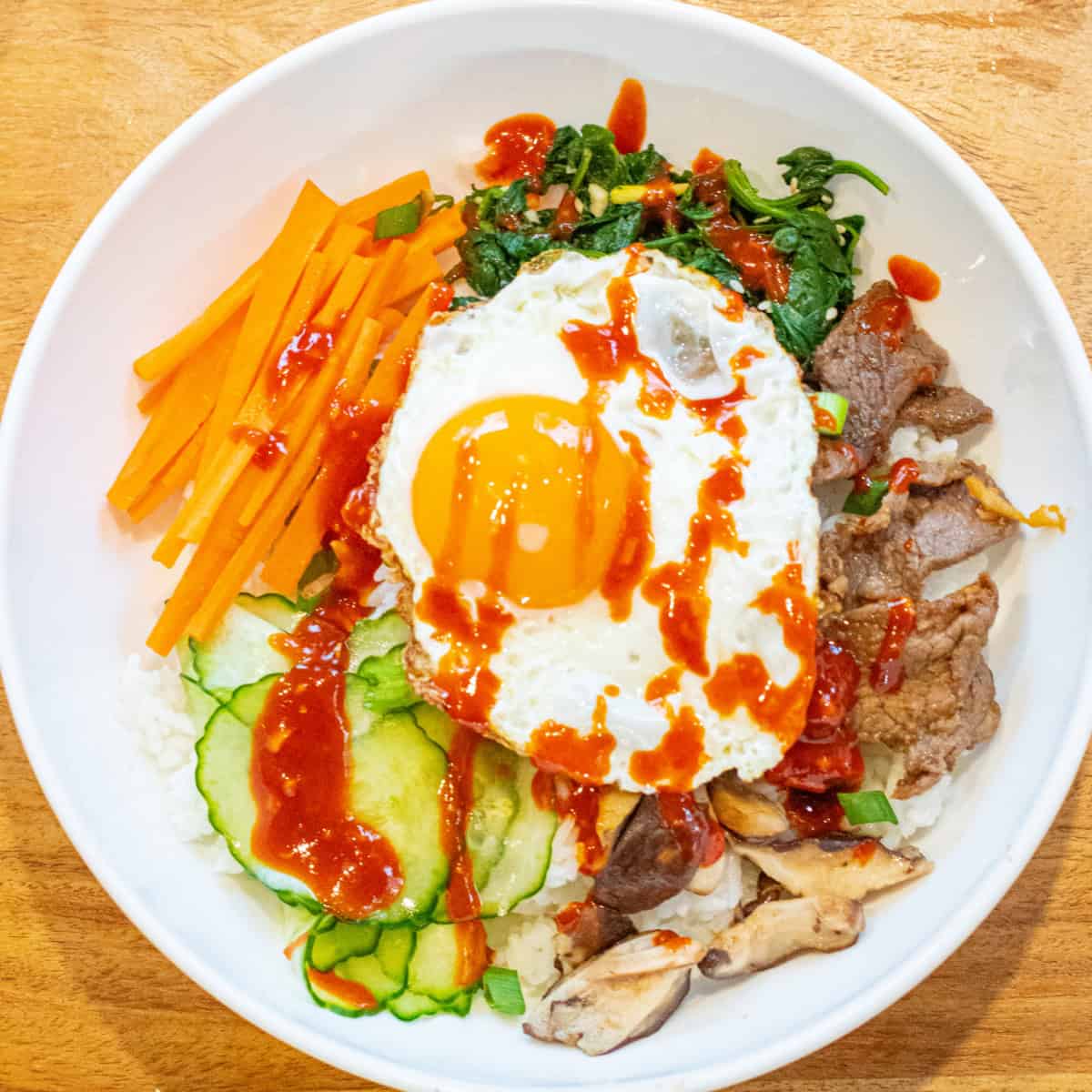 overhead of a big wide bowl of sliced carrots, cucumbers, spinach salad, beef, mushrooms, and an egg on top with sauce.