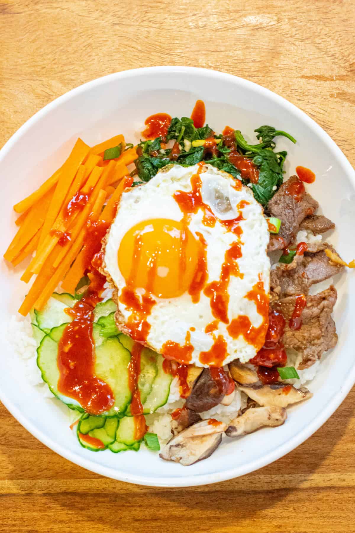 overhead of a big wide bowl of sliced carrots, cucumbers, spinach salad, beef, mushrooms, and an egg on top with sauce.