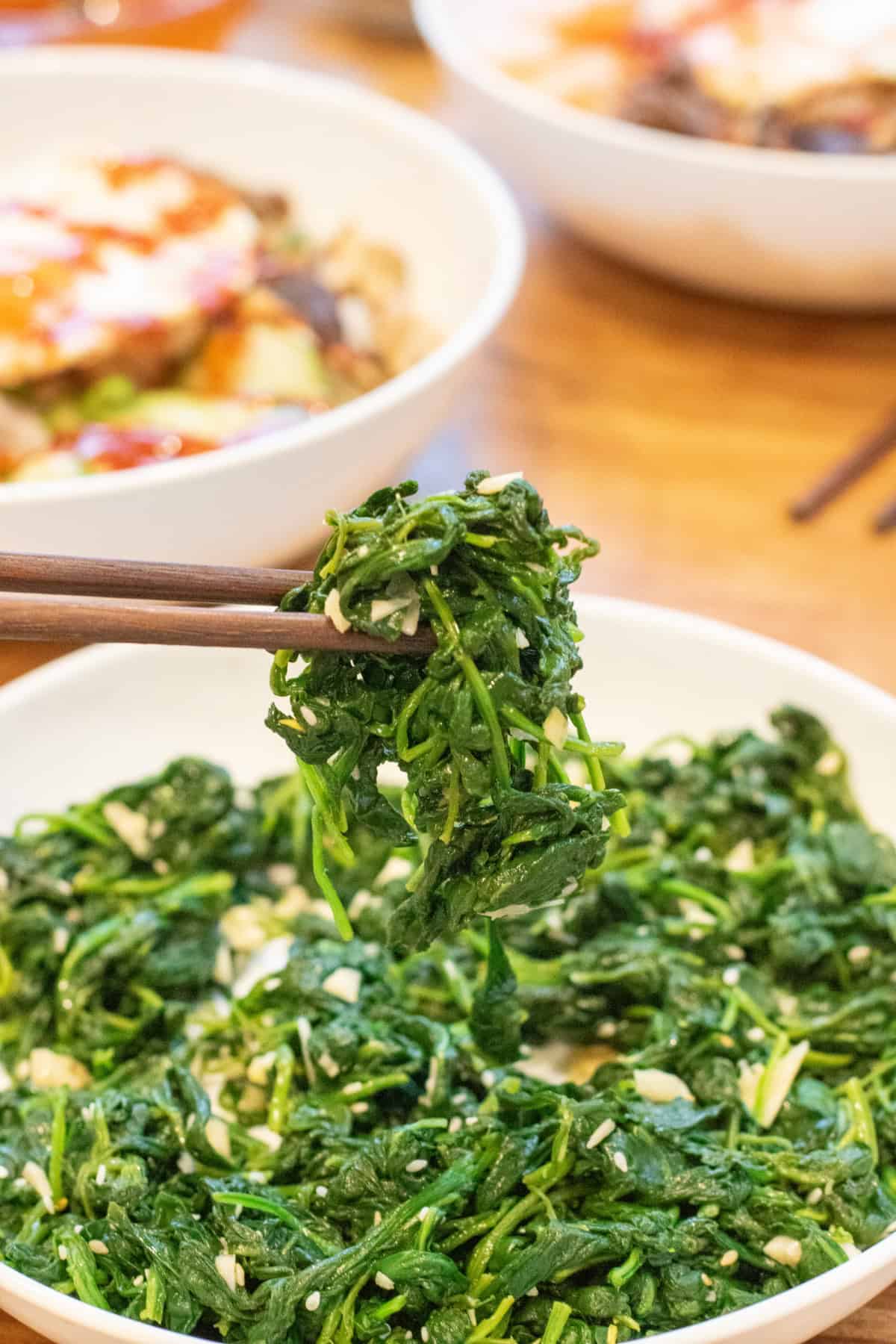 a bowl of blanched spinach with garlic and sesame seeds and some held up by chopsticks.