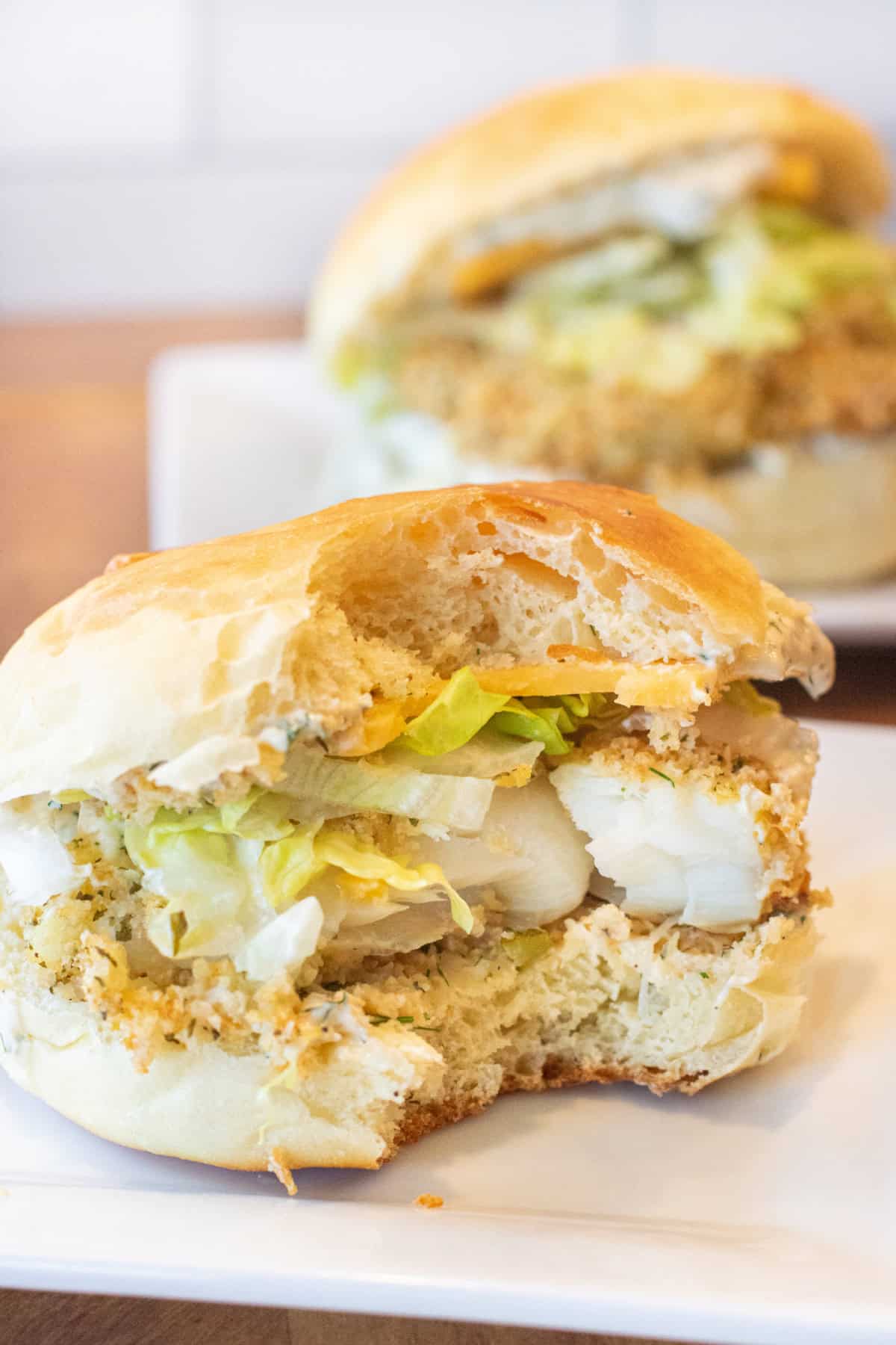 a breaded fish sandwich with lettuce and cheese on a brioche bun with a bite taken out and a sandwich on a plate behind it.