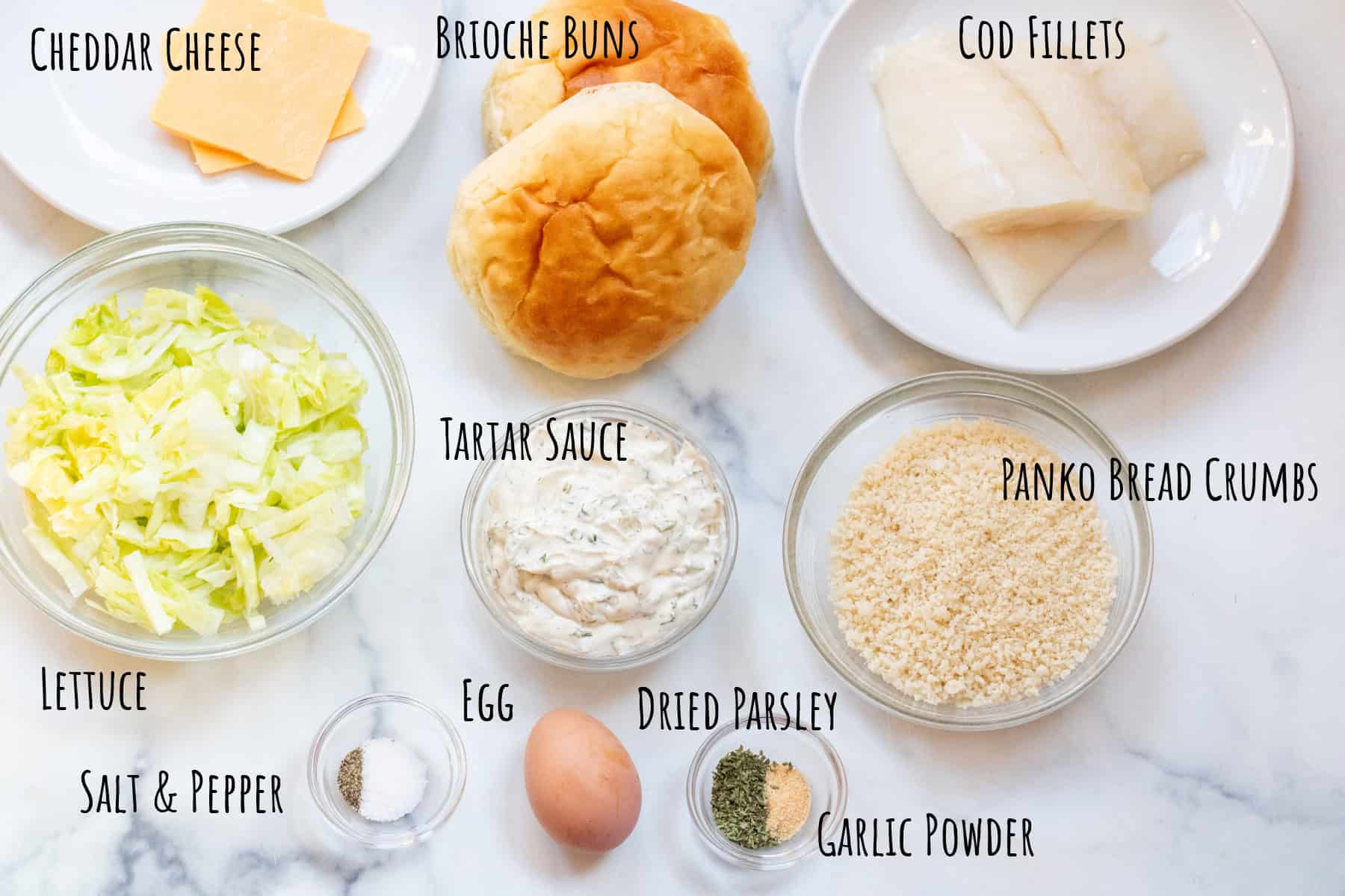 an overhead picture of slices of cheese, brioche buns, cod fillets, a bowl of shredded lettuce, tartar sauce, panko bread crumbs, egg, spices all labeled.